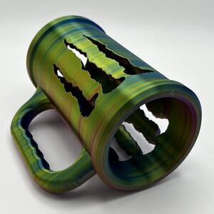 May include: A multi-colored mug with a unique design. The mug features a green, blue, and purple color scheme with a handle and cut-out claw marks. The mug is made of a durable material and is suitable for hot or cold beverages.