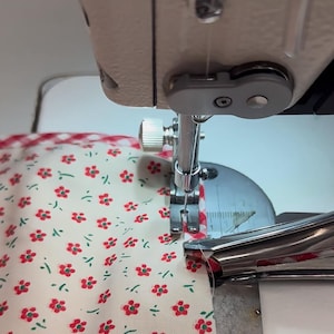 May include: Close-up of a sewing machine in action, stitching fabric. The fabric is white with a red floral pattern and a red and white gingham border. The needle is piercing the fabric, creating a seam.