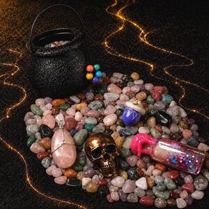 May include: A black cauldron filled with colorful stones sits atop a bed of assorted gemstones. A gold skull, a pink pendant, and a small bottle with glitter are also visible. The image has a mystical theme.