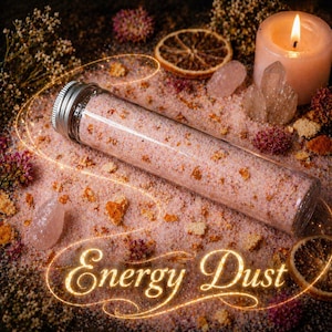 May include: A clear glass tube with a silver cap filled with pink and orange granules, labeled "Energy Dust". The tube rests on a bed of pink granules with dried flowers, crystals, and a lit candle. The background is dark.