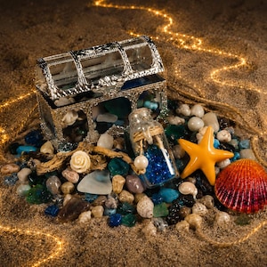 May include: A silver-toned treasure chest filled with assorted sea glass and small stones, accompanied by a glass bottle containing blue glass beads, a yellow starfish, a colorful seashell, and a small white rose, all arranged on sand.