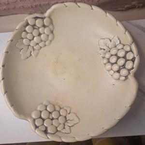 Pode incluir: Uma tigela redonda de cerâmica em tom branco sujo com três cachos de uvas em relevo e detalhes em cinza escuro nas bordas. A borda da tigela tem um design recortado com entalhes sutis e uma superfície com acabamento esmaltado liso.