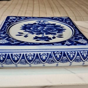 May include: A square ceramic tray featuring a white center with a blue floral illustration. The border displays a repeating blue and white geometric pattern, while the sides are decorated with a blue and white scalloped arch design.