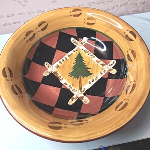 Puede incluir: Un cuenco decorativo de cerámica con un diseño rústico. El cuenco presenta un patrón de cuadros en el centro, un diamante con un árbol verde y un borde con huellas de pezuñas marrones. El borde es de color marrón claro.