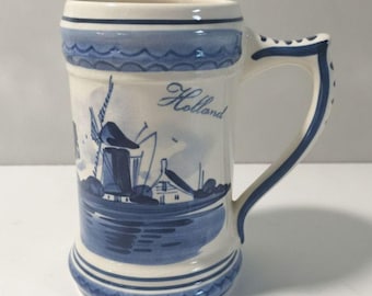 May include: A ceramic beer mug featuring a blue and white hand-painted design of a Dutch windmill and landscape. The word Holland is written in blue script on the front. The mug has a sturdy handle and decorative blue wave patterns at the rim and base.