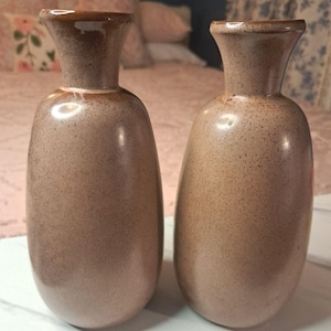 May include: Two matching ceramic vases with a smooth, speckled tan and beige glaze. Each vase features a rounded, bulbous body that tapers into a narrow neck with a flared rim. The finish has a subtle, mottled texture throughout the surface.