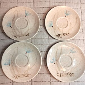 Può includere: Un set di quattro piattini rotondi in ceramica con finitura color crema maculata. Ogni piattino presenta un incavo circolare centrale e un disegno dipinto di semi di tarassaco azzurri sopra steli marroni e piccoli ciottoli.