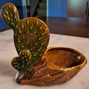 Może przedstawiać: Glazurowana ceramiczna doniczka w kształcie kaktusa opuncji przymocowana do teksturowanej brązowej podstawy. Kaktus jest oliwkowozielony z wypukłymi kolcami i posiada mały złoty emblemat w kształcie tarczy z napisem The Alamo oraz gwiazdką.