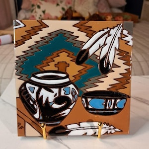 May include: A square ceramic tile featuring a painted scene of a black and white patterned pot and bowl on a brown surface. The background displays a geometric pattern in teal, brown, and white, accented by two white feathers with black tips.