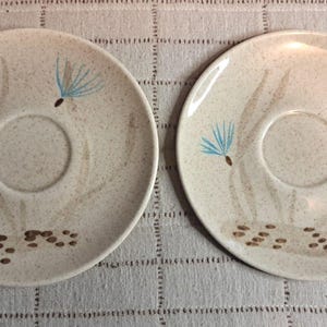 Puede incluir: Dos platillos redondos de cerámica con acabado beige moteado. Cada platillo presenta una hendidura circular central y un diseño pintado de guijarros marrones en la base con semillas de diente de león azul claro flotando arriba.