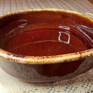 May include: A round ceramic baking dish with a glossy dark brown interior and exterior. The rim and the two small side handles feature a textured, mottled light tan and brown glaze finish, providing a distinct contrast to the smooth dark basin.