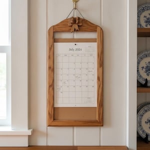 Pode incluir: Uma moldura de madeira para calendário de parede com um detalhe de laço esculpido no topo. A moldura contém um calendário de papel branco para julho de 2024, fixado por um alfinete. A madeira tem um acabamento de grão natural e um gancho.