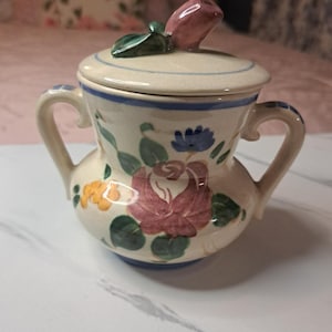 May include: A small, cream-colored ceramic sugar bowl with a lid, featuring hand-painted floral designs in pink, blue, yellow, and green. The lid is topped with a pink rose bud. The bowl has two handles and a blue trim.