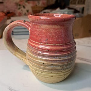 May include: A ceramic mug with a rounded body and a curved handle. The upper portion features a vibrant coral-pink glaze, while the lower section transitions into a soft cream color. The surface is decorated with horizontal ridges and dark speckles.