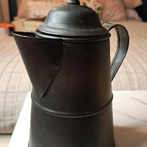 May include: A matte black metal coffee pot with a cylindrical body, a wide triangular spout, and a curved handle. The pot features a removable lid topped with a small, dark, tiered wooden knob. The surface has a dark, weathered finish.