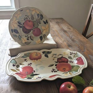 May include: A round plate and an oval platter featuring a painted pattern of red apples, green leaves, and dark berries on a cream background. Both pieces have scalloped edges and are displayed alongside two fresh red apples with green stems.