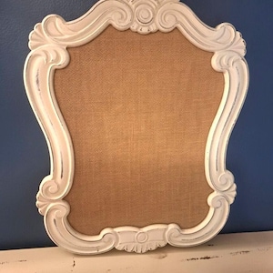 May include: A wall-mounted bulletin board featuring a tan burlap center panel framed by an ornate, white-painted wooden border with a distressed finish. The frame displays intricate scrollwork and carved shell-like details at the top and bottom.