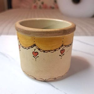 May include: A small, cylindrical ceramic cup with a beige base and a thick, unglazed brown rim. The exterior features a hand-painted yellow scalloped border near the top, accented with small, red heart motifs and delicate black line details.