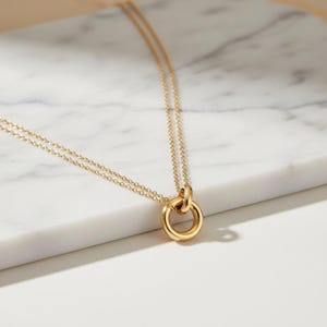 May include: A gold-colored necklace with a double chain and a circular pendant. The pendant features a smaller ring attached to a larger ring. The necklace is displayed on a white marble surface.