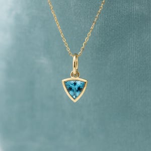 May include: A gold necklace featuring a triangular pendant with a faceted light blue gemstone. The pendant is suspended from a delicate gold chain, showcasing a polished metal setting that frames the vibrant blue stone in a simple, clean design.