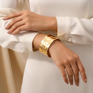 May include: A wide, gold-colored cuff bracelet with a hammered texture. The bracelet is worn on a wrist, and the image shows the bracelet's textured surface and the way it reflects light. The bracelet is a statement piece.