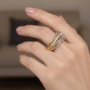 May include: A gold and silver ring with three interlocking bands. One band is silver, one is gold, and the third is gold with small, clear stones. The ring is worn on a finger.