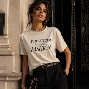 Puede incluir: Una camiseta color crema con el texto "TRAUMATIZED but make it FASHION" en negro. La camiseta se combina con pantalones negros y un cinturón negro con hebilla dorada. También se ve un bolso negro con cadena dorada.