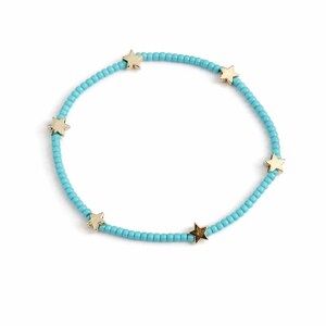 May include: A beaded bracelet featuring small, light blue beads and gold-colored star charms. The bracelet is displayed on a white background, showcasing its simple yet elegant design. The stars are evenly spaced around the bracelet.