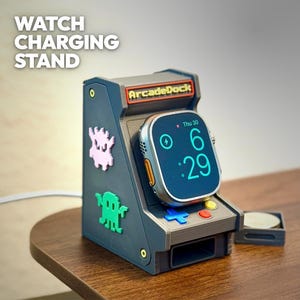 May include: A gray watch charging stand shaped like an arcade game, with the text "WATCH CHARGING STAND" and "ArcadeDock". The stand has colorful buttons and two monster-shaped stickers. An Apple Watch is docked, displaying the time.