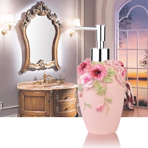 May include: A pink floral soap dispenser with a silver pump. The dispenser is decorated with pink and red flowers and green leaves. The dispenser is in a bathroom setting with a gold-framed mirror and a wooden vanity.