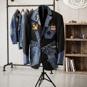 Könnte beinhalten: Ein schwarzer Blazer mit Jeans-Patches und Akzenten, präsentiert auf einem Stativ. Die Jacke hat Verzierungen mit Biene und Handtasche. Andere Jeansjacken hängen auf einem Gestell im Hintergrund. Ein einzigartiges Mode-Statement.