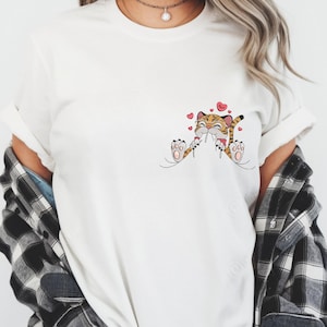 May include: White t-shirt with a cartoon tiger illustration. The tiger is orange and black, holding a flower with hearts. The design is on the front of the shirt. The tiger has a smiling face.