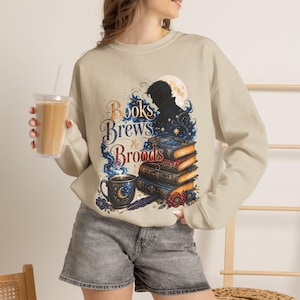 May include: A beige crewneck sweatshirt featuring a graphic print with the text "Books Brews & Broods". The design includes a stack of three books, a dark mug with a crescent moon, a silhouette of a person, and a full moon in the background.