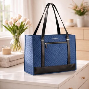 May include: A blue tote bag with a quilted pattern, black leather accents, and black handles. The bag has a zippered pocket and the word "EMERSON" printed on the front. The bag is sitting on a white surface.