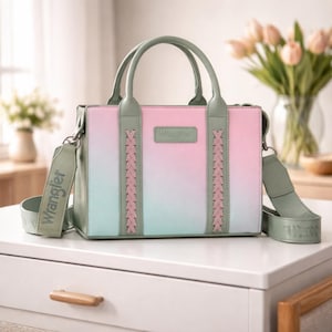 May include: A pastel-colored handbag with a gradient of pink, white, and teal. The bag has a top handle and a detachable shoulder strap. The brand name "Wrangler" is visible on the strap and the bag itself. Braided accents and a rectangular shape.