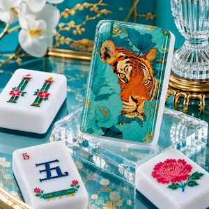 May include: A rectangular mahjong tile featuring a detailed orange tiger face emerging from teal lotus leaves. Other white tiles are present, including one with a red flower, one with a green bamboo design, and one with the Chinese character for five.