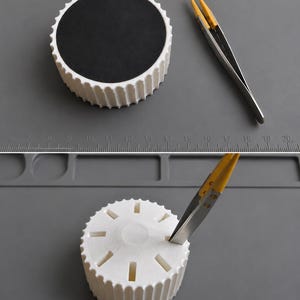 Pode incluir: Um suporte cilíndrico branco com exterior estriado e uma inserção circular preta no topo, acompanhado por pinças de metal com pegas amarelas. O suporte apresenta oito ranhuras retangulares na superfície superior para ferramentas.