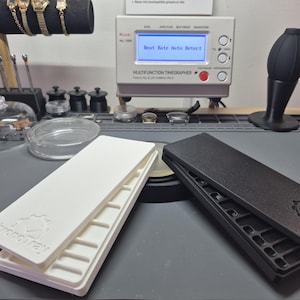 Può includere: Due vassoi per orologi, uno bianco e uno nero, sono aperti su una superficie grigia. Il vassoio bianco è impilato sopra il vassoio aperto. Il testo "ChronoTray" è visibile su entrambi i vassoi. Sullo sfondo c'è una macchina per la misurazione del tempo degli orologi.