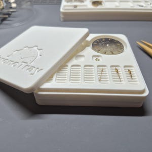 Może przedstawiać: Biały plastikowy organizer na części zegarków z napisem "ChronoTray" na pokrywie. Taca zawiera wskazówki zegarka, tarczę i inne małe elementy. Po prawej stronie widoczna jest pęseta.