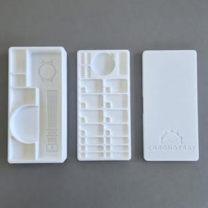 May include: A white watch organiser in three parts. The left section has a watch outline and strap design. The middle section has multiple compartments. The right section has the "CHRONOTRAY" logo. The organiser is designed to hold watches and watch accessories.