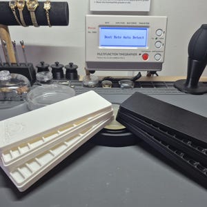 May include: A watch repair station setup with a multifunction timegrapher displaying "Beat Rate Auto Detect". Two stacks of watch trays, one white and one black, are in the foreground. Various watch tools and a watch display stand are also visible.