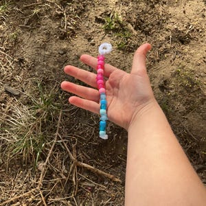 May include: A beaded keychain with a gradient of colors, transitioning from pink to blue. The keychain features a white loop at the top and is held against a natural, earthy background.