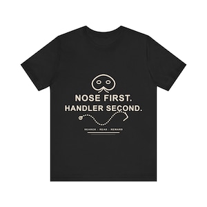 May include: A black short-sleeved t-shirt featuring a cream-colored graphic of a nose icon above the text NOSE FIRST. HANDLER SECOND. Below this, a dotted path line leads to an arrow, with the words SEARCH - READ - REWARD and a horizontal line.