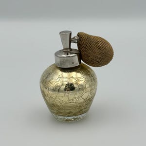 May include: A round perfume bottle with a gold crackle glass finish. The bottle features a silver-toned metal collar and a pump mechanism topped with a textured, brown fabric-covered bulb. The glass body has a distinct webbed pattern.
