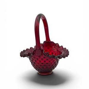 Op de afbeelding: Een dieprode glazen mand met een hobnail-textuur over het gehele buitenoppervlak. De mand heeft een hoog, gebogen handvat en een brede, geplooide, gekartelde rand. Het glas is doorschijnend, waardoor het licht door de levendige rode kleur valt.