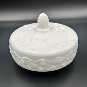 Puede incluir: Un plato blanco con tapa y un detallado diseño floral en relieve. La tapa presenta un pomo texturizado y los bordes del plato tienen un patrón festoneado. El diseño es de estilo clásico y vintage.