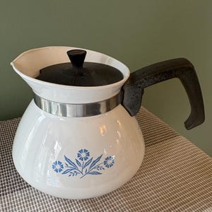 Puede incluir: Cafetera de esmalte blanco con tapa y asa negras. La cafetera presenta un diseño floral azul y una banda plateada. El asa es oscura y curvada, y el pico es ancho. Capacidad aproximada de 1,5 litros.