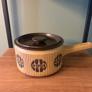 Op de afbeelding: Een beige keramische ovenschaal met een donkerbruin deksel en handvat. De schaal heeft een herhalend patroon van donkerbruine geometrische vormen. De ovenschaal staat op een houten oppervlak.