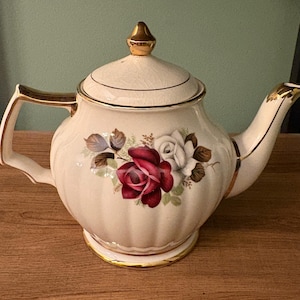 May include: Cream-coloured ceramic teapot with gold-coloured handle, spout, and accents. The teapot features a floral design with red and white roses. The lid is topped with a gold finial. The teapot is a vintage style.