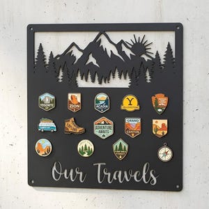 May include: Black metal wall art with a mountain and forest silhouette, holding travel-themed pins. The pins feature national park and adventure designs, with the text "Our Travels" at the bottom.